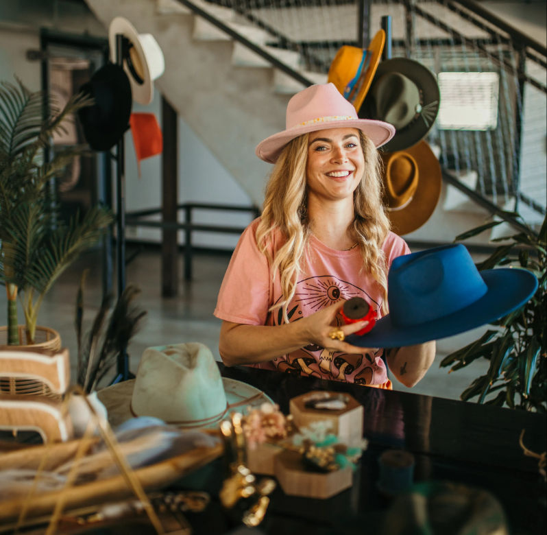 Smiling woman wearing a pink hat and graphic tee in a bright artisan hat studio, holding a blue felt fedora amid colorful hats, tools, and display racks.