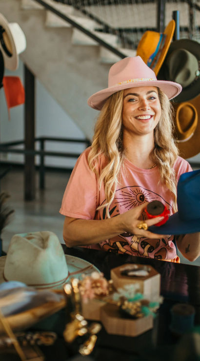 Smiling woman in a pink hat and tee shaping a bright blue fedora at an artisan hat studio, surrounded by hat racks, tools, and potted plants