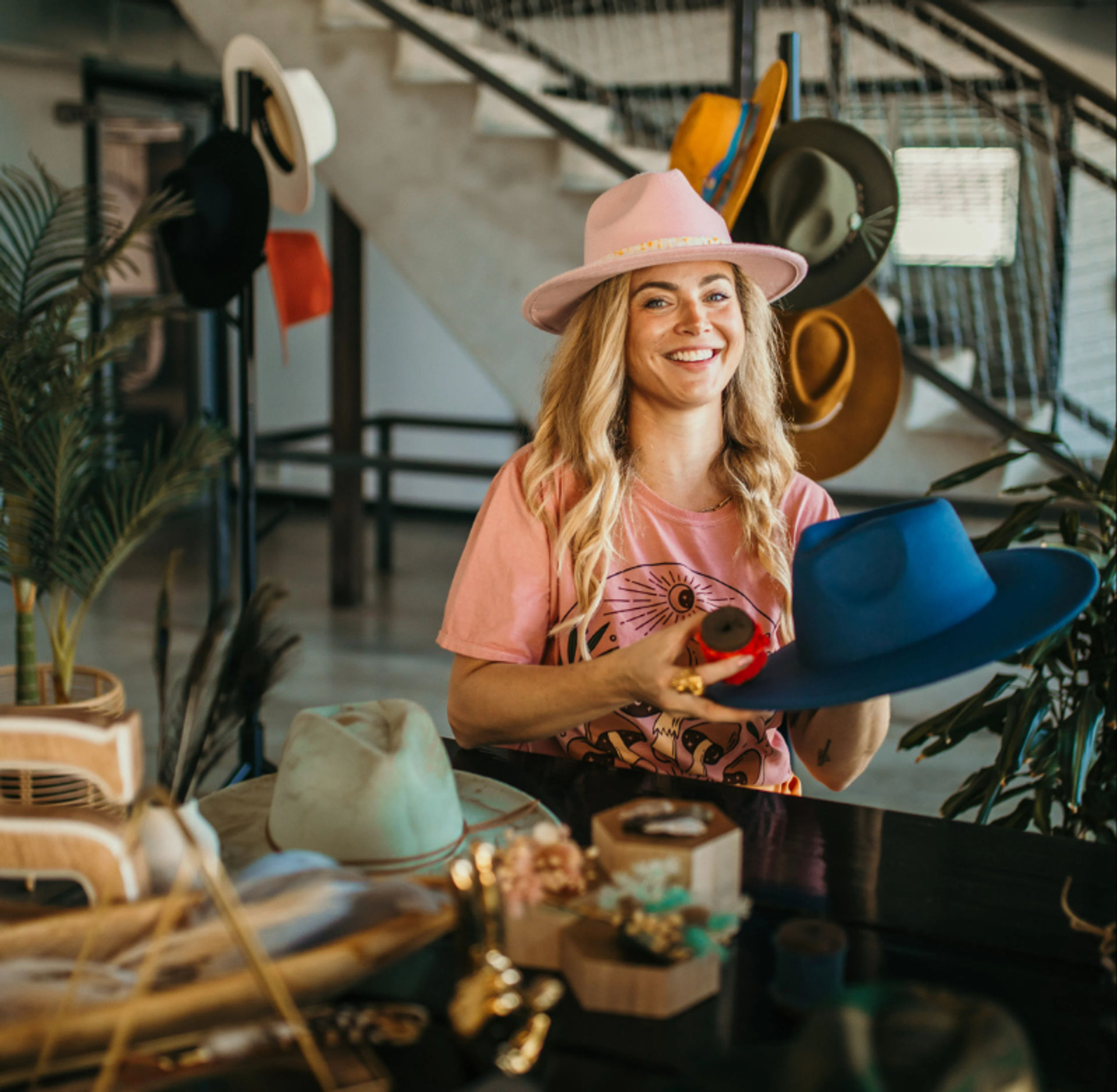 Smiling woman in a pink hat and tee shaping a bright blue fedora at an artisan hat studio, surrounded by hat racks, tools, and potted plants