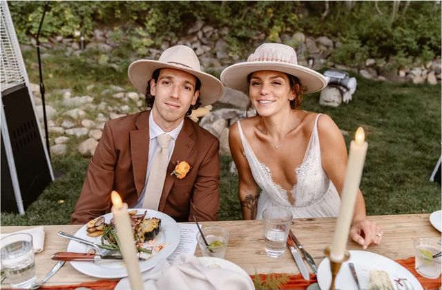 Smiling bride and groom in wide-brim hats at an outdoor rustic wedding reception on a lawn, seated at a wooden table with lit taper candles, plated dinner and a stone-and-greenery backdrop