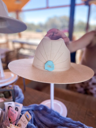 Beige wide-brim felt cowboy hat with a turquoise stone accent and stitched sunburst detail, displayed on a stand at an outdoor market stall.