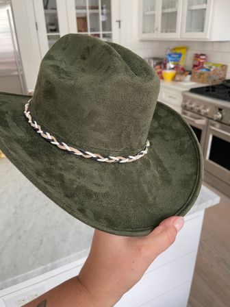 Olive-green suede cowboy hat with braided tan-black-white band held over a modern white kitchen with stainless steel stove