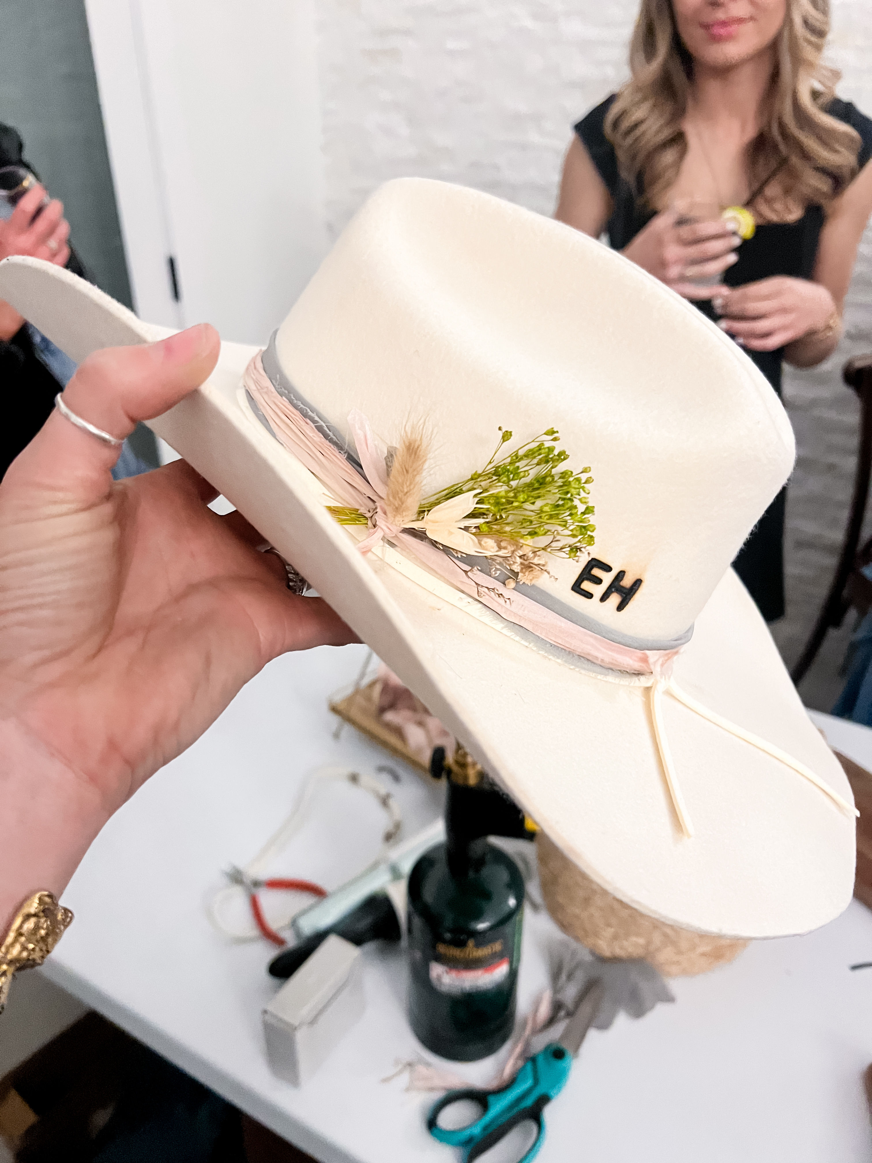 White felt cowboy hat held over a craft table, trimmed with pink-gray ribbon, dried floral sprig and 'EH' monogram, surrounded by scissors and tools.