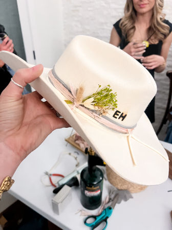 White felt cowboy hat held over a craft table, trimmed with pink-gray ribbon, dried floral sprig and 'EH' monogram, surrounded by scissors and tools.