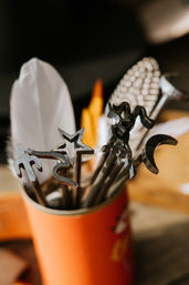 Close-up of metal shape stamps—star, crescent, squiggle and more—nestled with feathers in an orange tin, craft tools on a wooden studio workspace.
