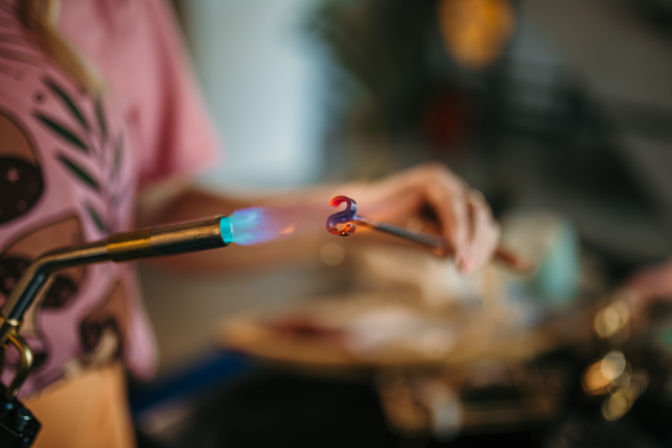 Close-up of a blue torch flame heating a small molten glass spiral on a rod in a lampworking studio as hands shape the glowing glass