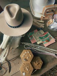 Southwestern-style flat lay: cream wide-brimmed felt hat with black stitched band on a rustic wooden table alongside a bleached cow skull, colorful bandanas, sparkly necklaces and a hexagonal jewelry display of earrings and charms — boho western accessories.