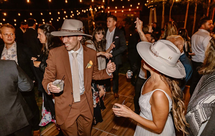 Couple in wide-brim hats dancing on a wooden dance floor at an outdoor nighttime wedding reception, man in a brown suit holding a cocktail and woman in a white dress with a drink, string lights and guests in the background.