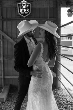 Smiling bride in lace gown and groom in suit wearing cowboy hats embrace in a rustic barn beneath a star-shaped neon sign.