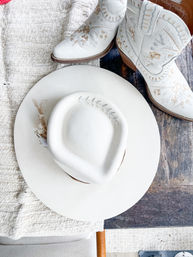 White felt cowboy hat with stitched leaf and dried floral accent on a rustic wooden farmhouse table beside embroidered white leather cowboy boots — Western boho accessory flatlay