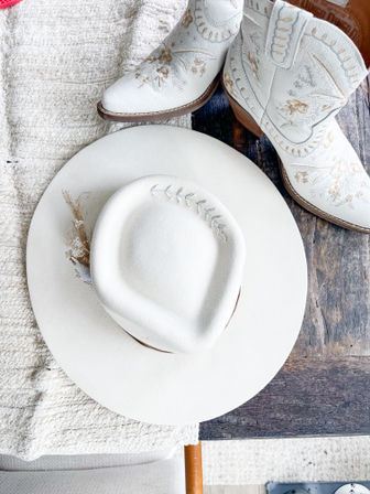 White felt cowboy hat with stitched leaf and dried floral accent on a rustic wooden farmhouse table beside embroidered white leather cowboy boots — Western boho accessory flatlay