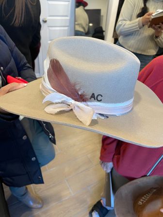 Beige wide-brim felt fedora with frayed ribbon bow and brown feather accent, displayed indoors in a boutique-style shop among shoppers.