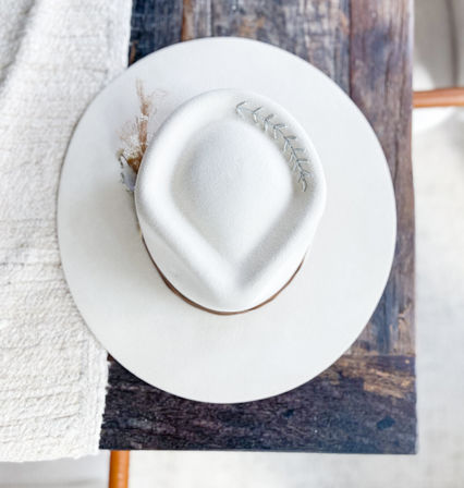 Top view of a white felt fedora with a brown band and small silver leaf pin, resting on a rustic wooden table next to a textured cream blanket