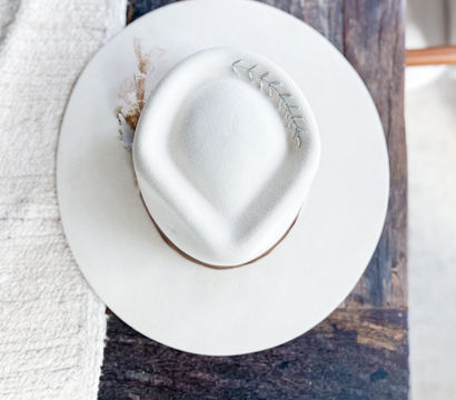Top view of a white felt fedora with a brown band and small silver leaf pin, resting on a rustic wooden table next to a textured cream blanket