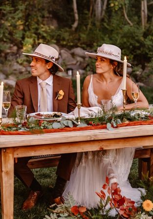 Boho bride and groom wearing wide-brim hats seated at a candlelit wooden table during a rustic outdoor wedding, lace gown and brown suit with boutonniere, greenery runner and champagne glasses.