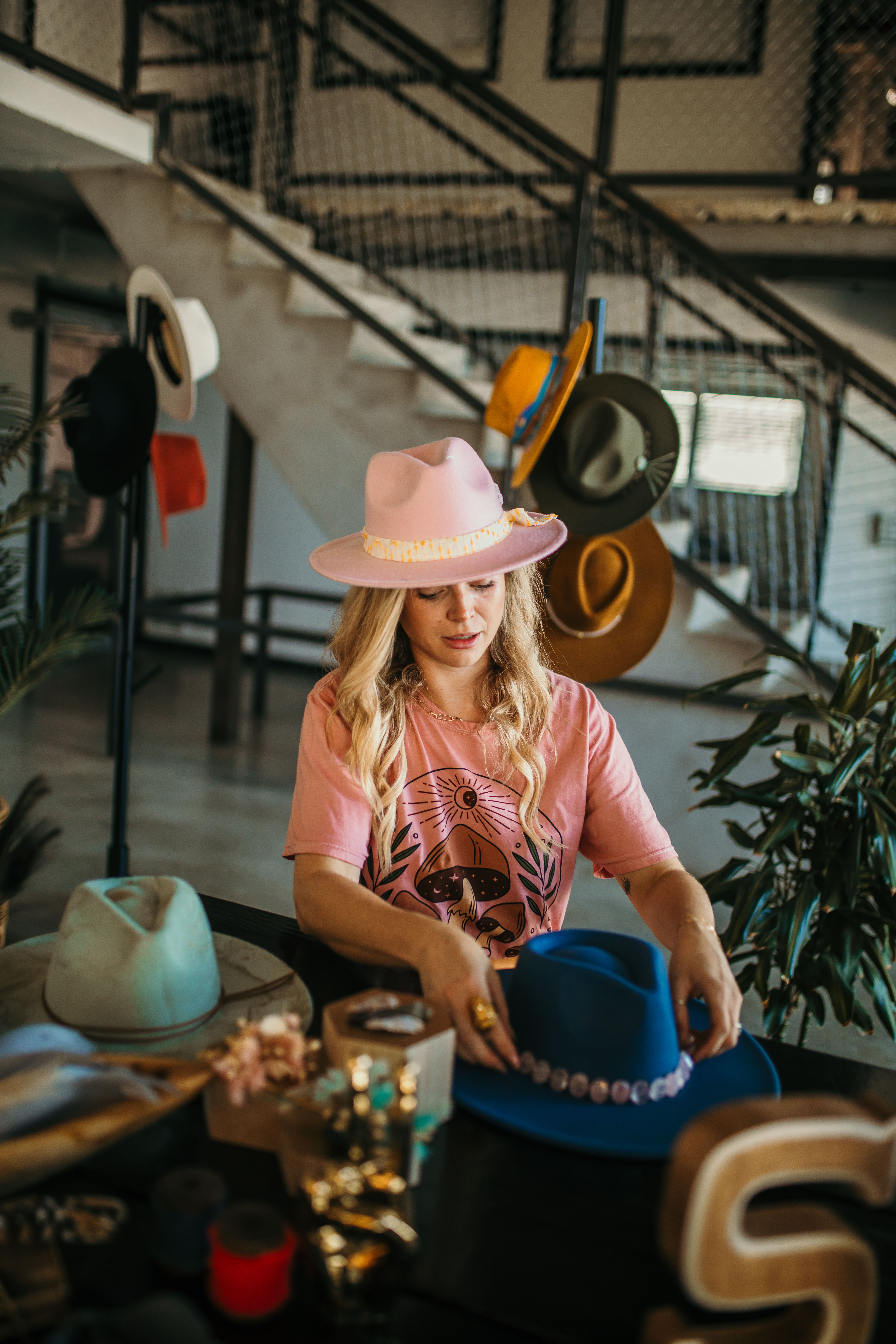 Blonde woman wearing a pink fedora arranging colorful felt hats and decorative hatbands on a table in a stylish hat shop with an industrial staircase backdrop