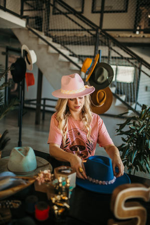 Blonde woman wearing a pink fedora arranging colorful felt hats and decorative hatbands on a table in a stylish hat shop with an industrial staircase backdrop