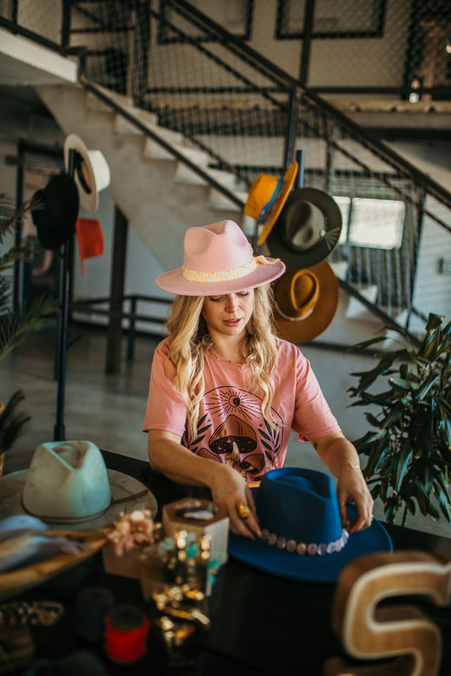 Blonde woman wearing a pink fedora arranging colorful felt hats and decorative hatbands on a table in a stylish hat shop with an industrial staircase backdrop