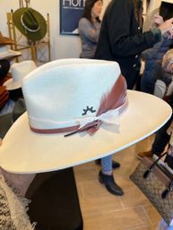 Cream felt wide-brim fedora with dusty-rose ribbon and feather and small sunburst logo, held in a hat boutique with shoppers in the background