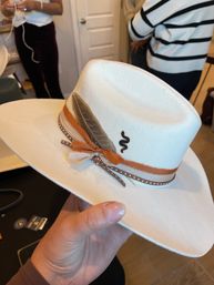 Hand holding a white felt Western cowboy hat with brown leather and orange ribbon, studded trim, feather accent and small black squiggle embroidery, indoor shop background