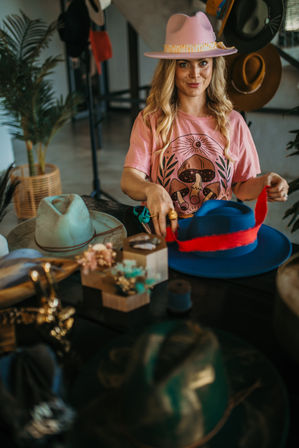 Woman in a boho millinery studio crafting a blue wide-brim hat with a red band, wearing a pink mushroom graphic tee and pink hat