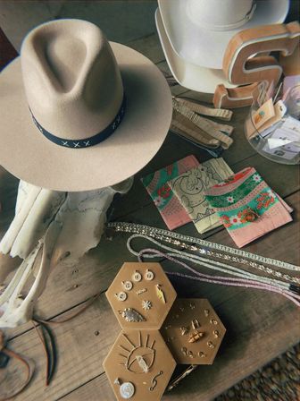 Beige wide-brim fedora with a black stitched band on a rustic wooden table, surrounded by a bleached skull prop, colorful printed bandanas, sparkling necklaces and hexagonal trays of gold and crystal earrings — Southwestern-style accessory display.