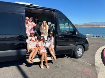 Smiling group of women in summer outfits posing in the open sliding door of a black passenger van at a sunny lakeside parking area with mountains and a boat dock in the background