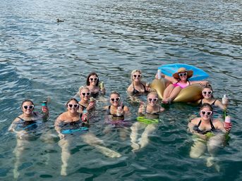 Group of friends wearing heart-shaped sunglasses floating in a lake, holding canned drinks and tumblers, one relaxing on a gold inflatable tube with a sun hat — fun summer lake day.