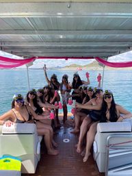 Group of friends in captain hats and black swimsuits toasting with pink can koozies on a decorated pontoon boat during a sunny lake boat party with rocky shoreline in the background.
