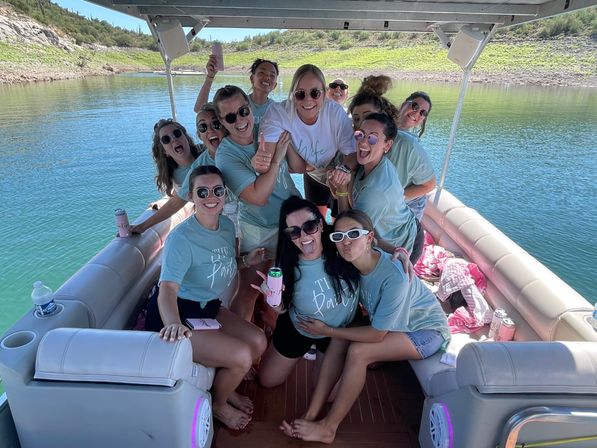 Smiling group of women in matching teal shirts and one in white celebrating on a pontoon boat, holding drinks on a sunny lake with rocky, green shoreline.