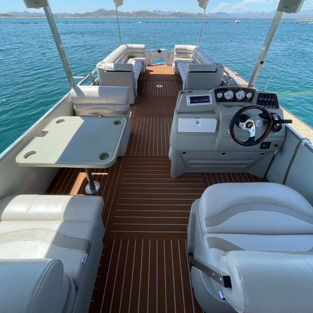 Pontoon boat interior with brown faux-teak deck, white cushioned seating, captain’s helm and small table, floating on clear turquoise water with a distant mountain shoreline on a sunny day.