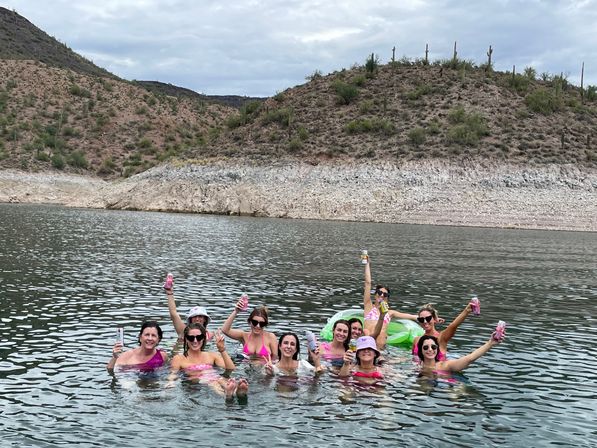 Group of friends floating and cheering with canned drinks in a desert lake, pink swimwear and a green inflatable, rocky shoreline and saguaro-studded hills under a cloudy sky.