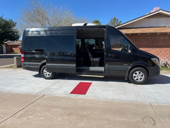 High-roof black passenger van with open sliding door parked on a suburban driveway beside a brick house, red welcome mat on the concrete.