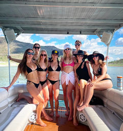 Eight women in swimsuits and sun hats smiling and posing on a covered pontoon boat deck on a sunny lake day with rocky hills and blue sky in the background.