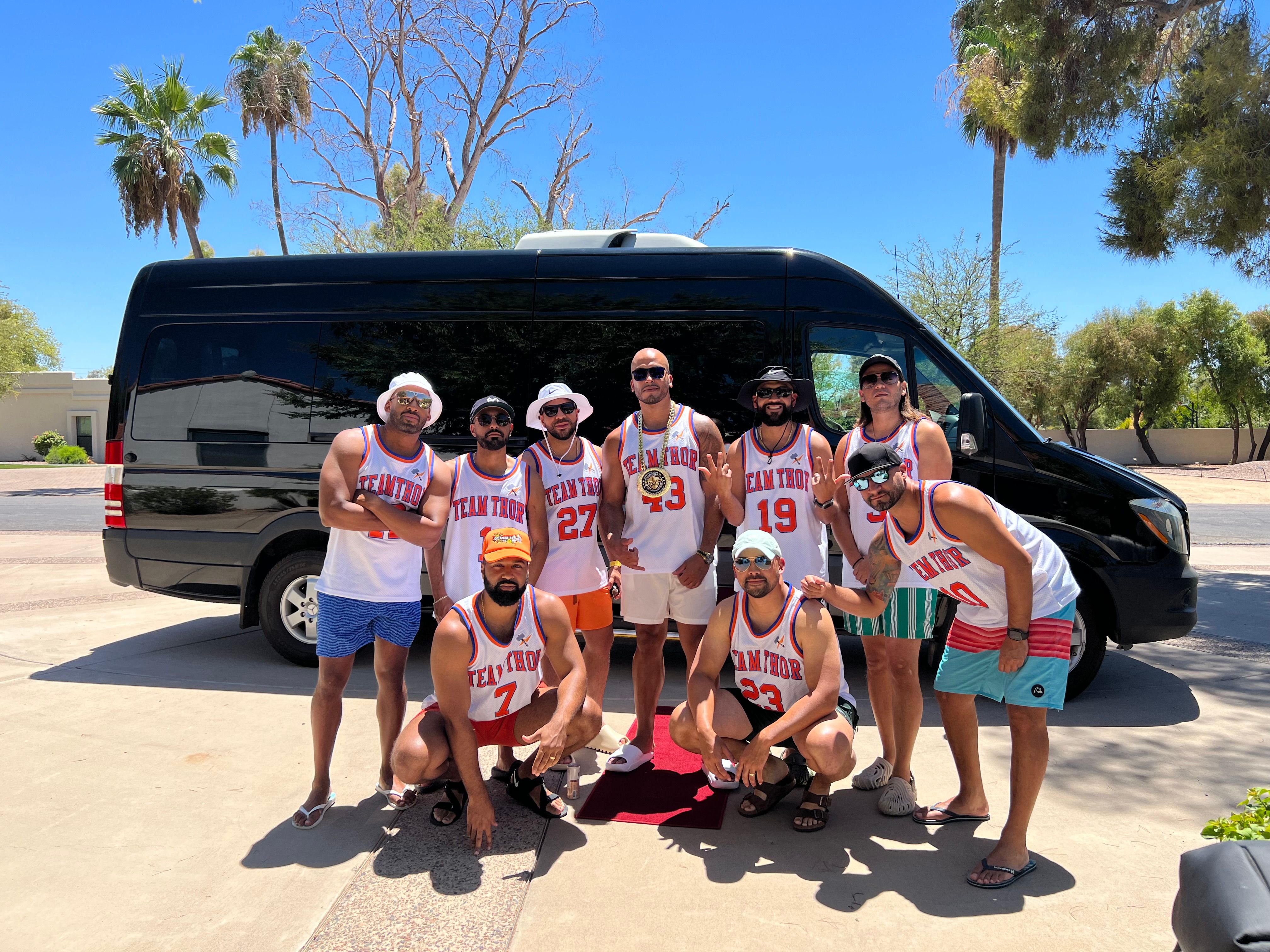 Ten men in matching "Team Thor" basketball jerseys posing in front of a black passenger van on a sunny suburban street with palm trees and bright blue sky.