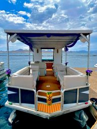 Pontoon boat moored at a scenic lake dock with distant mountains, shaded white canopy, white leather lounge seating, teak-style deck, purple floral accents and a tiki mask on the bow under a partly cloudy blue sky.
