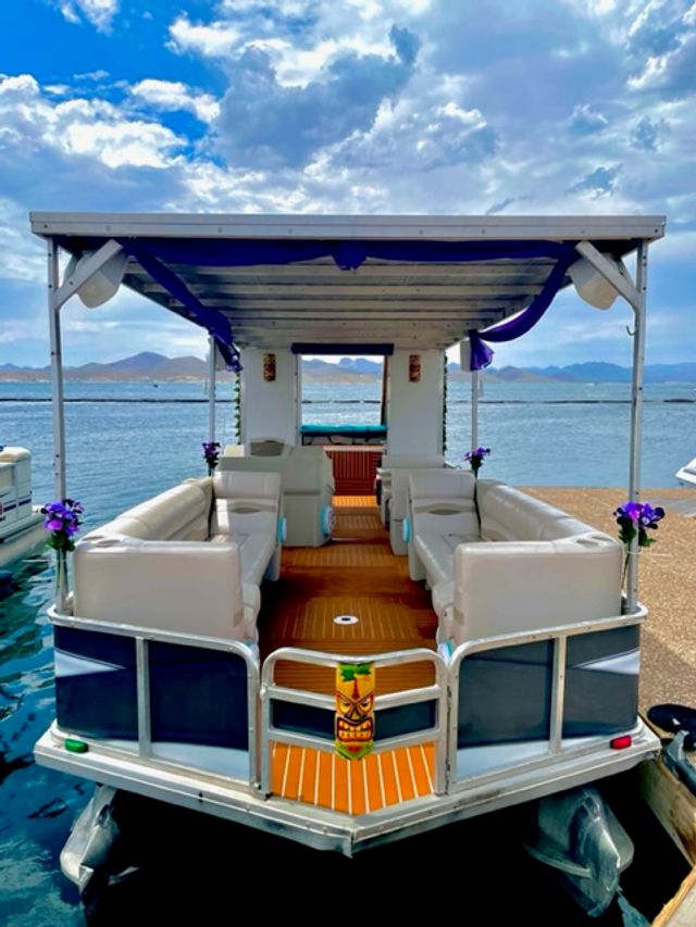 Pontoon boat moored at a scenic lake dock with distant mountains, shaded white canopy, white leather lounge seating, teak-style deck, purple floral accents and a tiki mask on the bow under a partly cloudy blue sky.