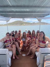 Smiling group of women in matching 'babe' swimsuits and a bride in white on a pontoon boat, enjoying a sunny bachelorette boat party on a desert lake with cactus-covered hills in the background.