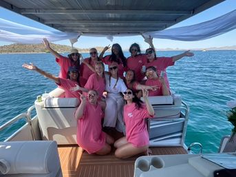 Group of women in matching pink shirts with one in white posing and cheering on a covered pontoon boat on a sunny blue lake with low hills in the background