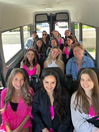 Smiling group of women in bright pink outfits posing inside a party shuttle minibus — lively girls’ day out or bachelorette party with sunglasses and big smiles.