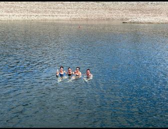 Four friends floating together in a blue lake near a rocky shoreline, smiling and holding drinks on a sunny day.