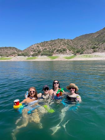 Four friends floating in a turquoise desert lake near rocky, cactus-covered hills under a clear blue sky, smiling and holding drinks in inflatable cup holders.