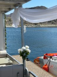 White sheer drape and bouquet of white flowers on a pontoon dock overlooking a blue lake, rocky shoreline, and an orange powerboat