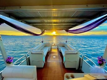 Pontoon boat interior at sunset with white cushioned benches, purple draped fabric and floral accents, open view over calm lake waters toward a distant mountain horizon.
