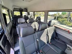 Sunlit passenger van interior with multiple rows of black leather seats, seatbelts, and large windows showing a palm-lined suburban street.