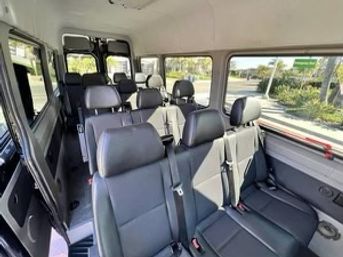 Sunlit passenger van interior with multiple rows of black leather seats, seatbelts, and large windows showing a palm-lined suburban street.