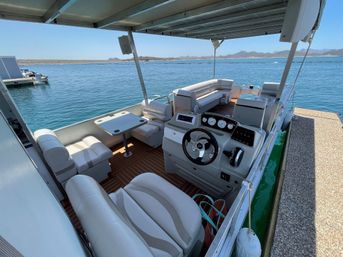 Spacious pontoon boat interior with white cushioned seating, helm with steering wheel and gauges, small table and covered deck docked on a clear blue lake with distant hilly shoreline on a sunny day