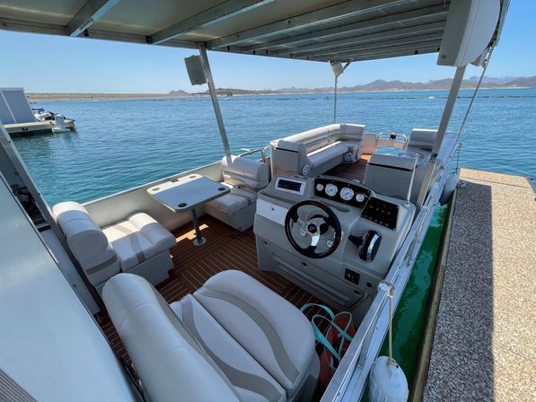 Spacious pontoon boat interior with white cushioned seating, helm with steering wheel and gauges, small table and covered deck docked on a clear blue lake with distant hilly shoreline on a sunny day