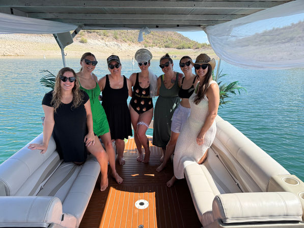 Seven smiling women in swimsuits, sundresses, sunglasses and sun hats posing together on a covered pontoon boat over blue-green lake water with a rocky shoreline in the background.
