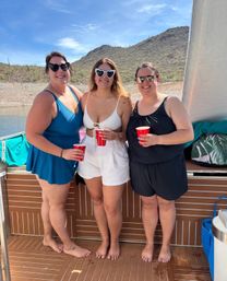 Three friends on a boat at a desert lake, wearing swimsuits and sunglasses and holding red cups, smiling in front of rocky, cactus-covered hills under a bright blue sky.